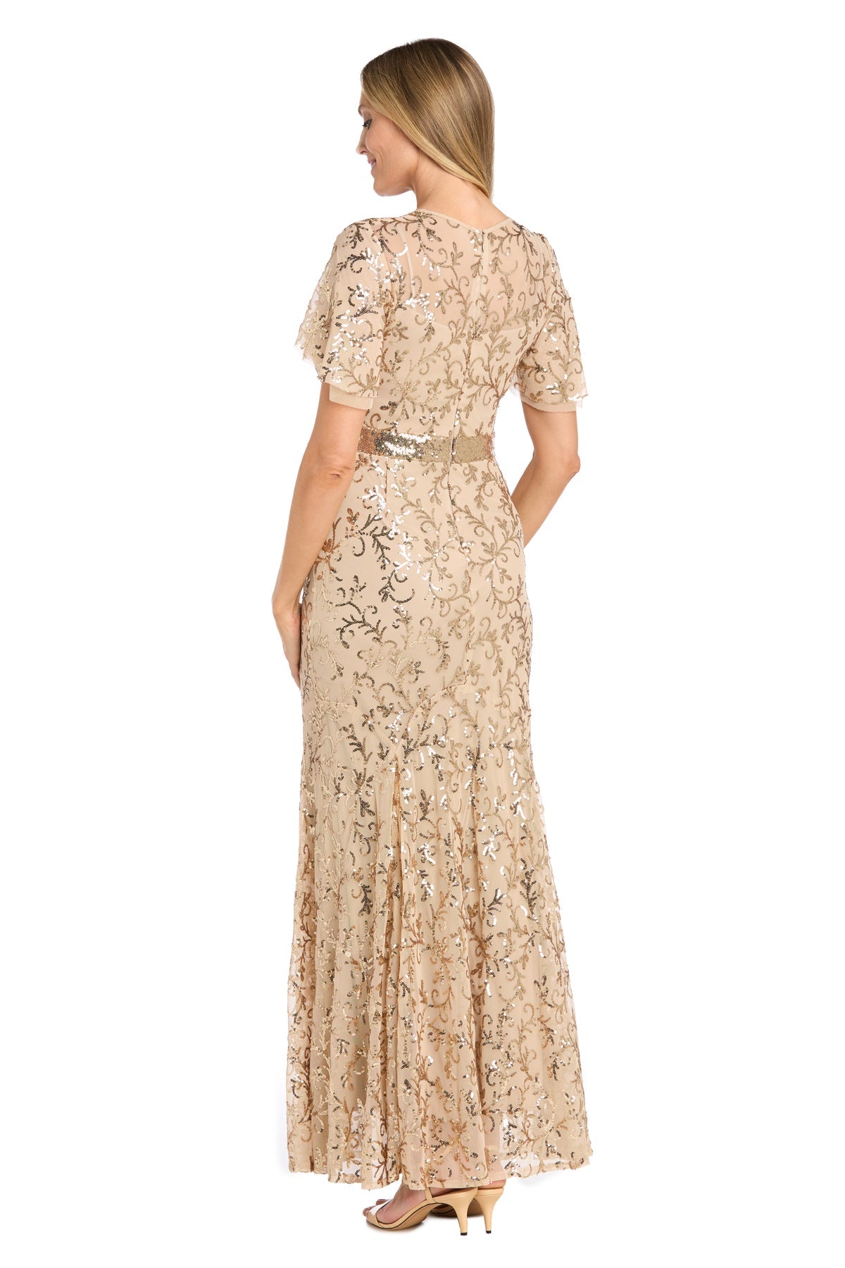 A woman stands facing away, wearing the R&M Richards 3425 Long Gold Sequin Formal Dress with heeled sandals.